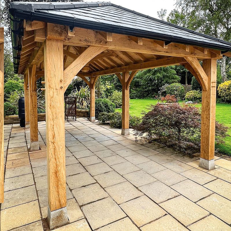 Open wooden gazebo with thick timber posts and curved braces on a stone-paved patio, surrounded by a green garden with shrubs and trees.