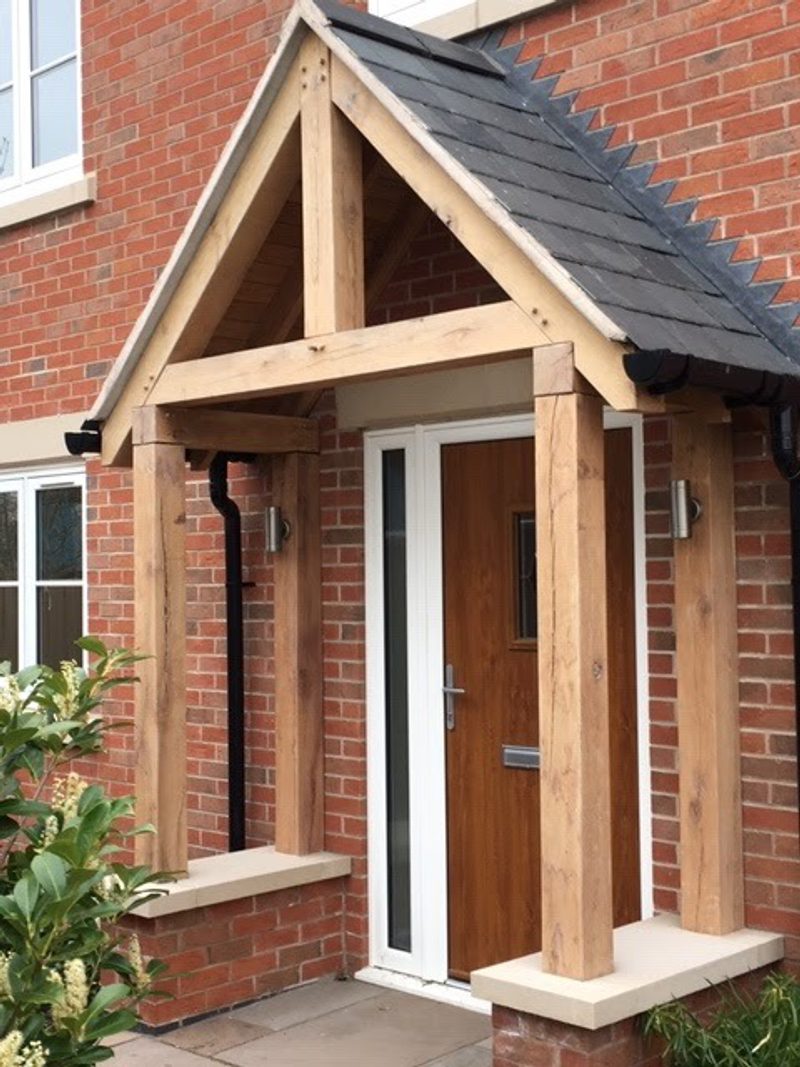 Brick house entrance with a timber-framed porch, slate roof, and a wooden door with white trim; plants nearby.