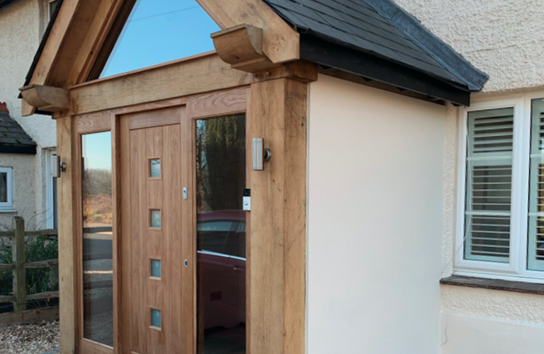 Wood-framed entrance with a triangular roof on a white house; central wooden door with square windows, side glass panels, and brick base.