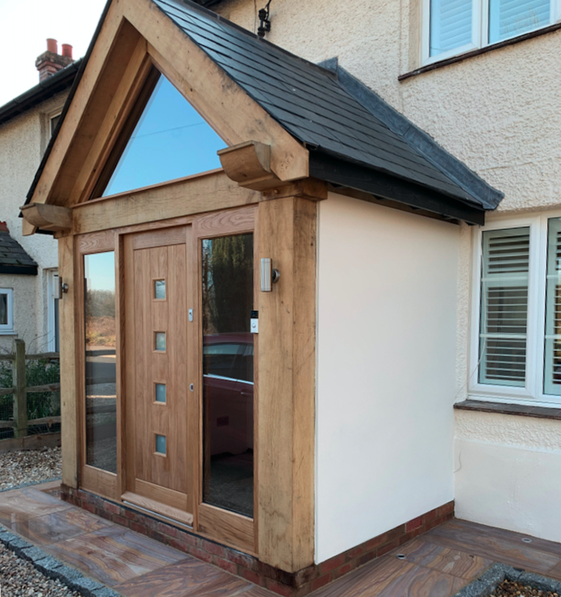 Wood-framed entrance with a triangular roof on a white house; central wooden door with square windows, side glass panels, and brick base.