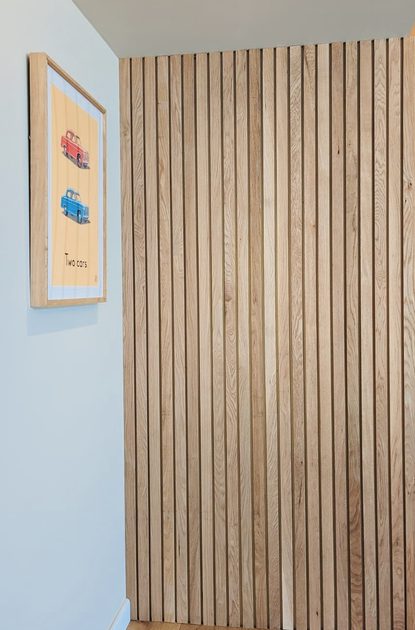 Corner of a room: vertical wooden slat wall on the right, light blue wall with a framed poster of two cars (red and blue), ceiling light, wooden floor.