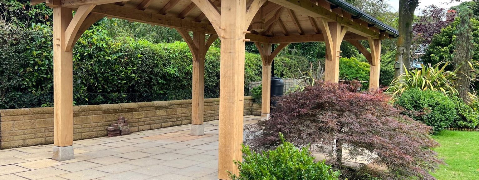 Wooden gazebo with a dark tiled roof on a paved patio, supported by curved posts, surrounded by hedges and trees.