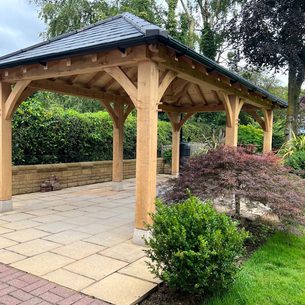 Wooden gazebo with a dark tiled roof on a paved patio, supported by curved posts, surrounded by hedges and trees.