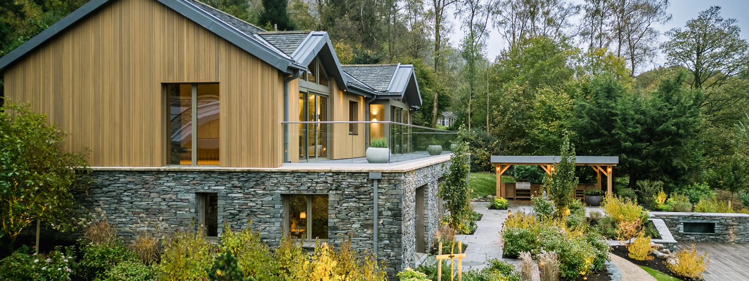 Two-story house with wood siding over a stone base, glass-railed balcony, and a landscaped garden with a curved gravel path and a wooden pergola in a wooded yard.