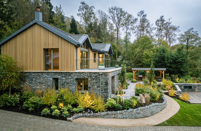 Modern two-story home with a stone base, timber siding, glass balcony, and a curved landscaped garden with stone walls and a wooden pergola.