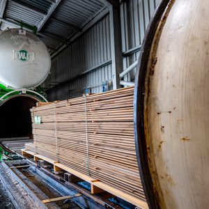 Industrial lumber-drying facility: a green circular kiln with a large door, stacked wooden planks on a pallet beside metal walls and a concrete floor.