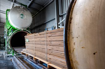 Industrial lumber-drying facility: a green circular kiln with a large door, stacked wooden planks on a pallet beside metal walls and a concrete floor.