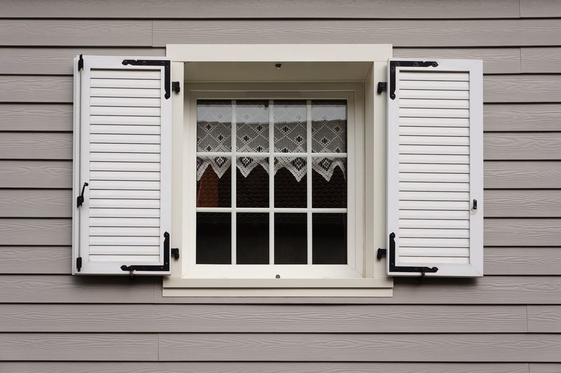 White-framed window with a 4x3 pane grid and lace curtains, open white shutters on beige horizontal siding, with a reflection of a red-tiled roof in the glass.