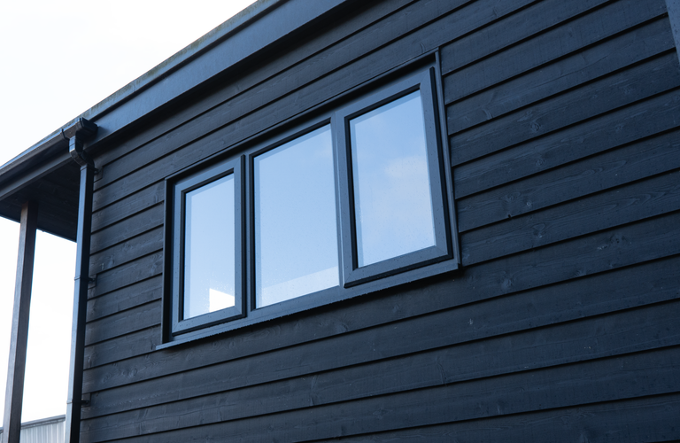 Angled view of a dark blue wooden exterior with three rectangular windows framed in black; gutter and roof edge visible on the left.