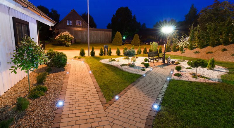 Night garden with two illuminated brick paths leading toward a central circular gravel bed with a bench and small trees, and a bright lamp in the background.