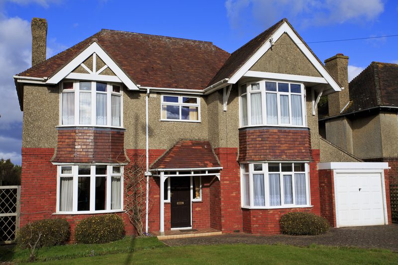 Two-story house with pebbledash and red brick accents, white-framed bay windows, a small front porch over the dark door, and a white garage on the right under a blue sky.