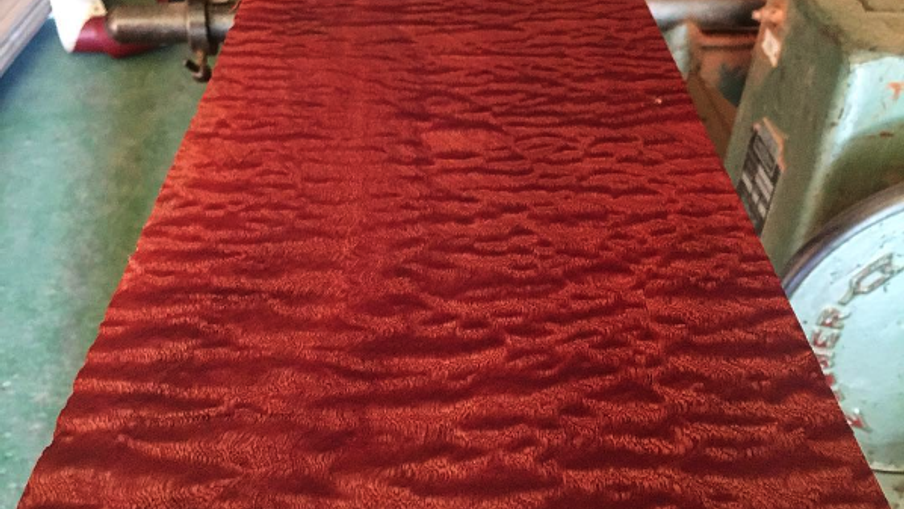 A long red quilted wood panel with a wavy figure extends along a workshop bench; green machinery and wooden pallets are visible in the background.