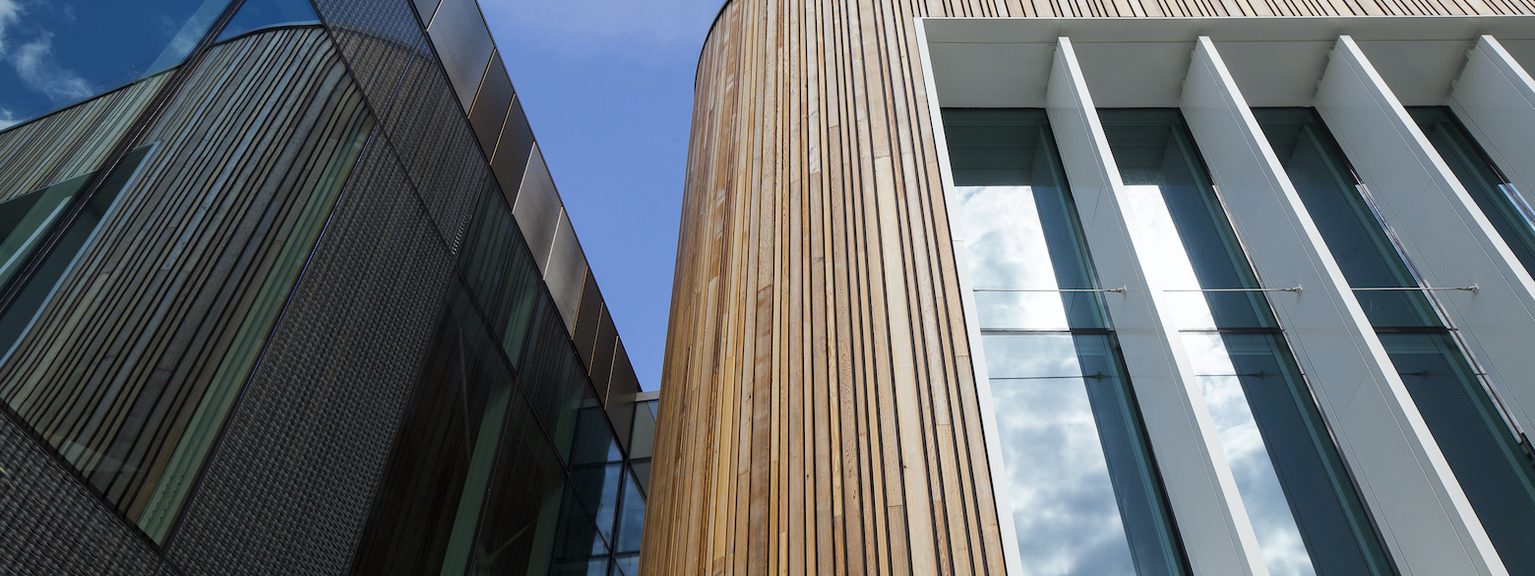 Modern building facade with vertical wooden slats on a curved corner, large white-framed glass windows, and a dark glass wall reflecting the blue sky.