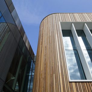 Modern building facade with vertical wooden slats on a curved corner, large white-framed glass windows, and a dark glass wall reflecting the blue sky.