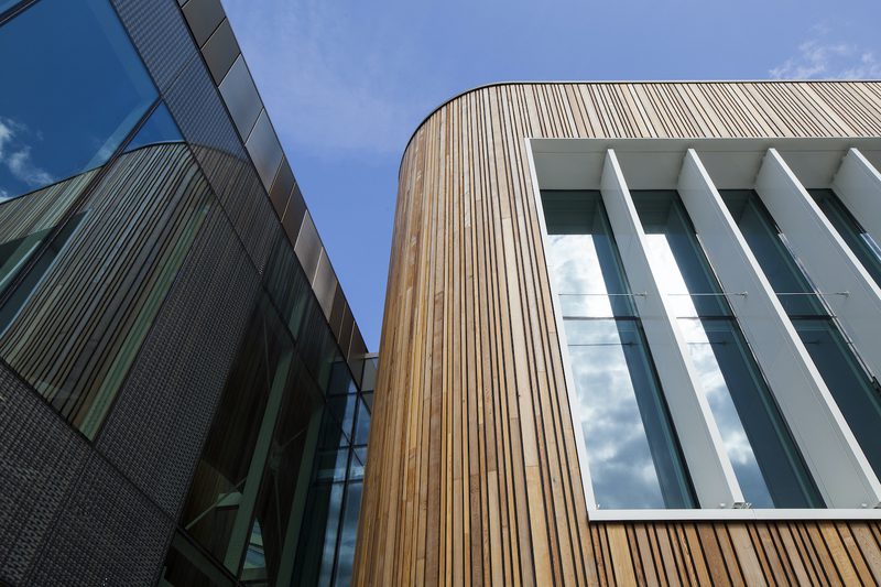 Modern building facade with vertical wooden slats on a curved corner, large white-framed glass windows, and a dark glass wall reflecting the blue sky.