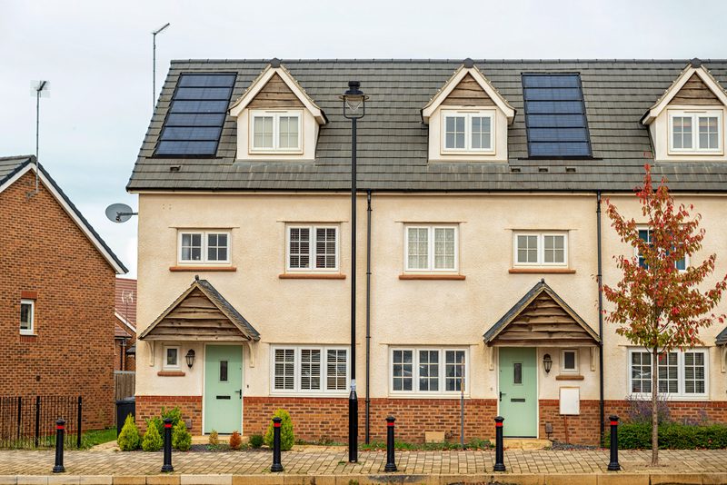 Row of cream stucco townhouses with brick bases and pastel green doors, two dormer windows on a tiled roof with solar panels, a lamp post, and a tree.