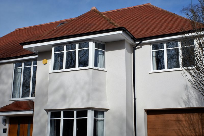 Two-story white house with a red-tiled roof, bay windows, and a wooden garage door; yellow exterior light, leafless tree, and blue sky.