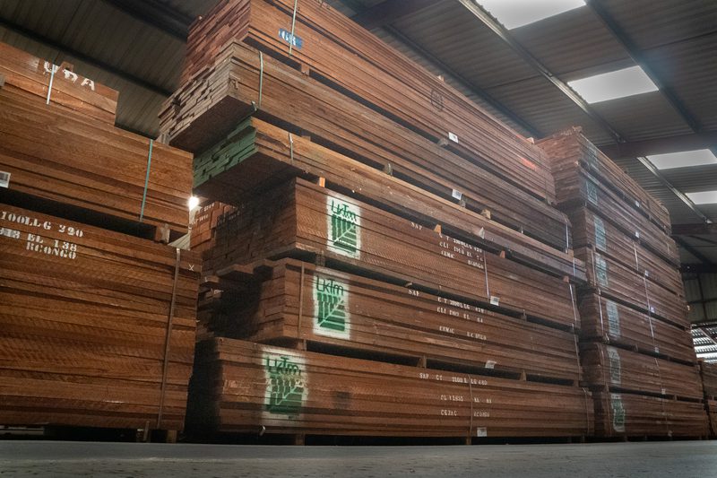 Industrial warehouse with tall stacks of brown wooden boards bound in bundles, green labels on the sides, and bright overhead lights.