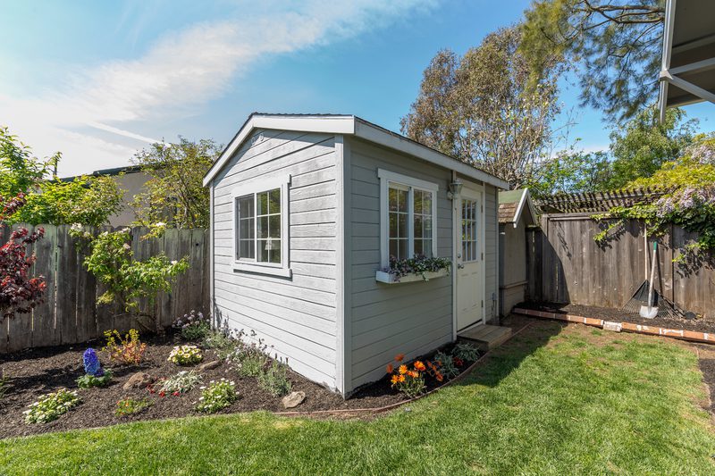 Small gray shed with white trim in a sunny backyard; window with a flower box, a door, garden beds along a wooden fence, and a rake nearby.