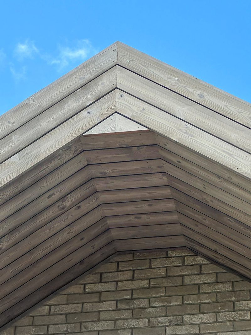 Low-angle view of a layered wooden gable roof above a brick wall, set against a clear blue sky.