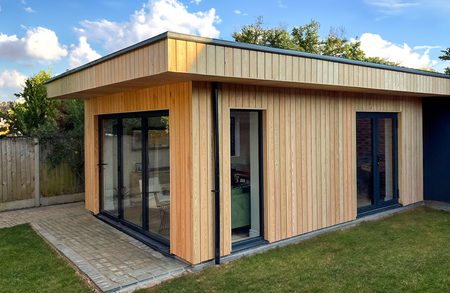 Modern wooden-clad single-story extension with large glass doors and a flat roof, set on a paved patio beside a green lawn.