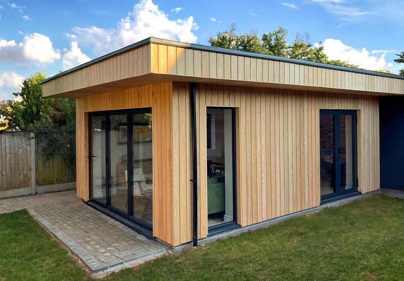 Modern wooden-clad single-story extension with large glass doors and a flat roof, set on a paved patio beside a green lawn.