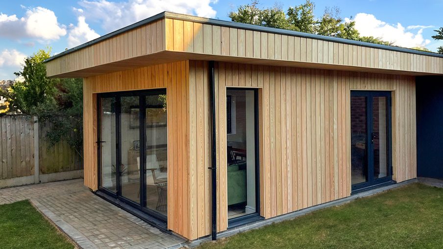 Modern wooden-clad single-story extension with large glass doors and a flat roof, set on a paved patio beside a green lawn.