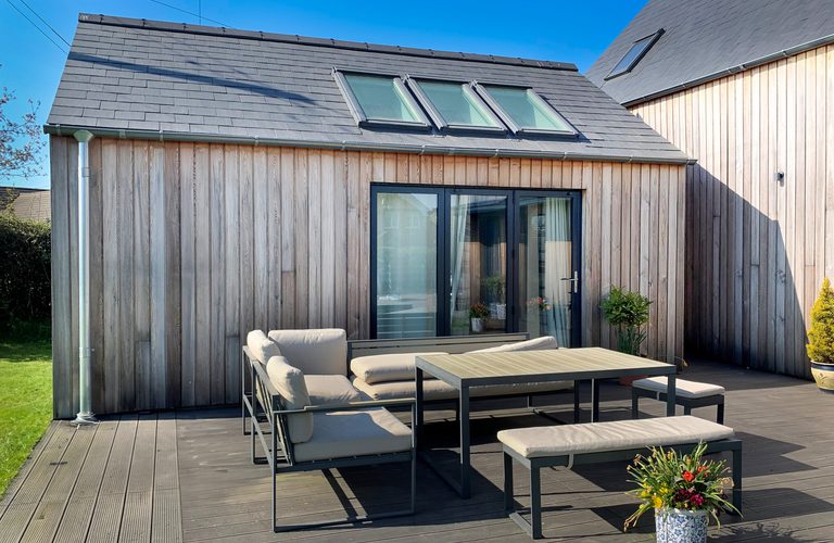 Modern wooden house with slate roof and skylights, glass doors, and a gray cushioned outdoor dining set on a wooden deck with plants and blue sky.