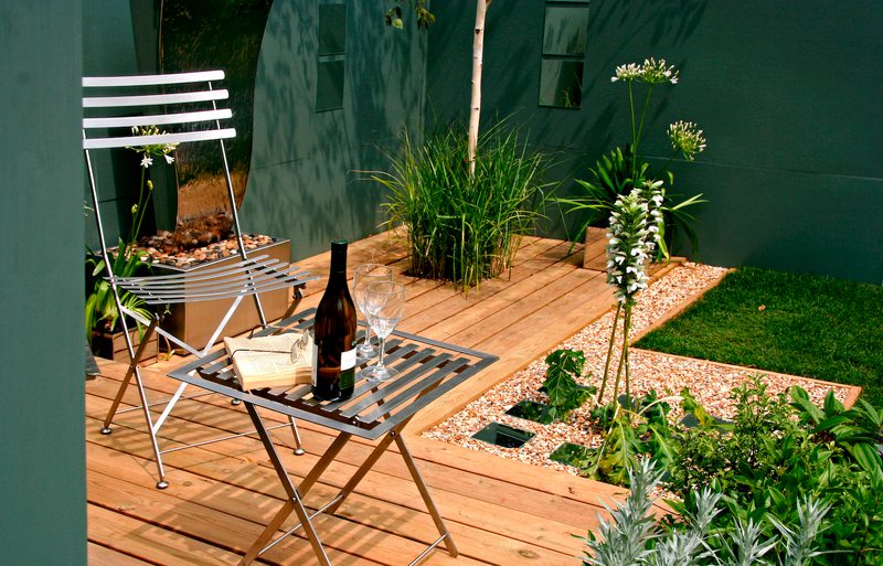 Wooden patio with a white metal bistro chair and small table holding a wine bottle, two glasses, and an open book; gravel paths, tall grasses, and green walls.