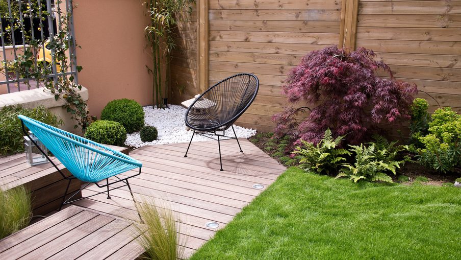 Wood deck garden with turquoise string chair, black round chair, green lawn, ferns, purple shrub, and white pebbles along a wooden fence.
