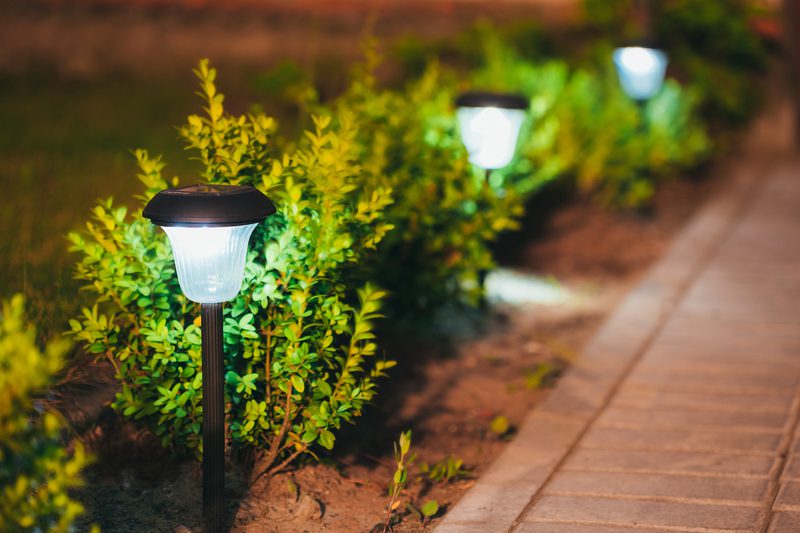Nighttime scene of a brick-paved sidewalk, with warm-glowing garden lamps along a leafy shrub border.