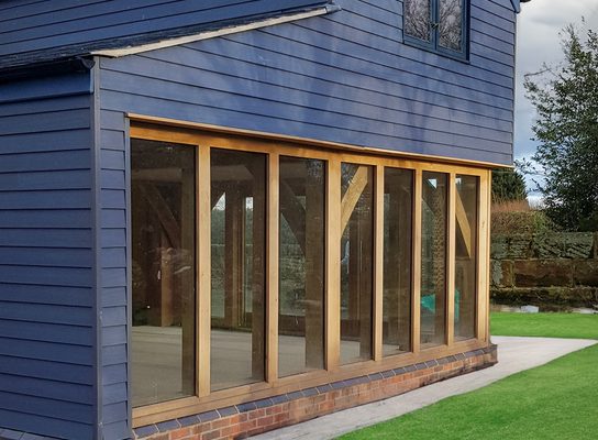 Blue house with a gabled roof and a glass-walled extension framed in wood, set on a brick base beside a green lawn under a cloudy sky.