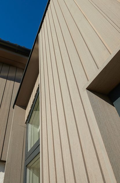 Angled view of a modern beige building with vertical siding and a window, set against a clear blue sky.