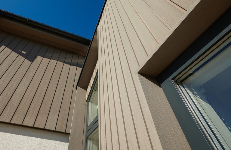 Angled view of a modern beige building with vertical siding and a window, set against a clear blue sky.