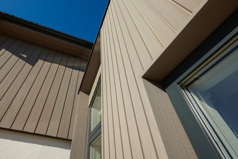 Angled view of a modern beige building with vertical siding and a window, set against a clear blue sky.