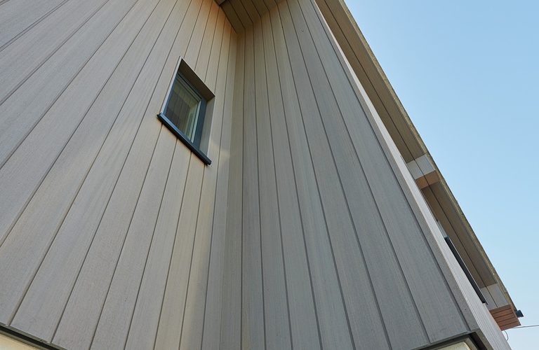 Low-angle view of a modern house corner with vertical gray siding, a small square window, a white stucco base, and a clear blue sky.