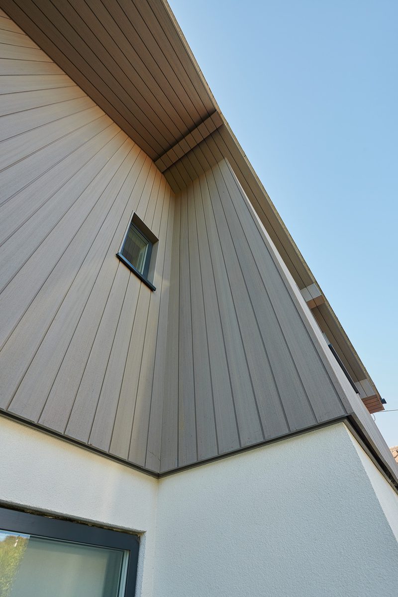 Low-angle view of a modern house corner with vertical gray siding, a small square window, a white stucco base, and a clear blue sky.