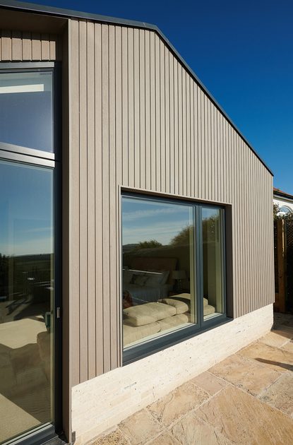 Exterior of a modern house with vertical gray-brown siding, a large glass door and window, a light stone base, and a stone patio beneath a clear blue sky.