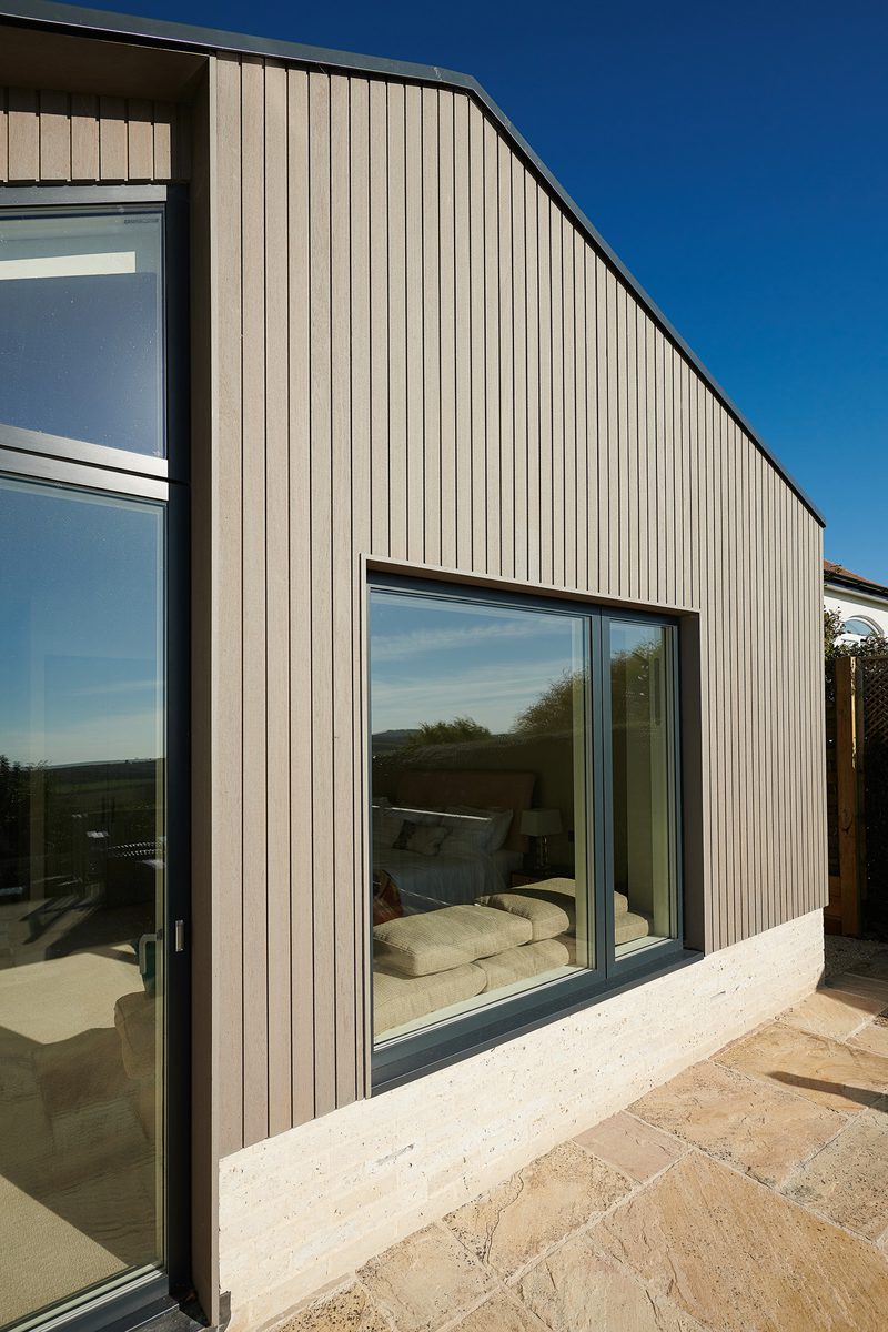 Exterior of a modern house with vertical gray-brown siding, a large glass door and window, a light stone base, and a stone patio beneath a clear blue sky.