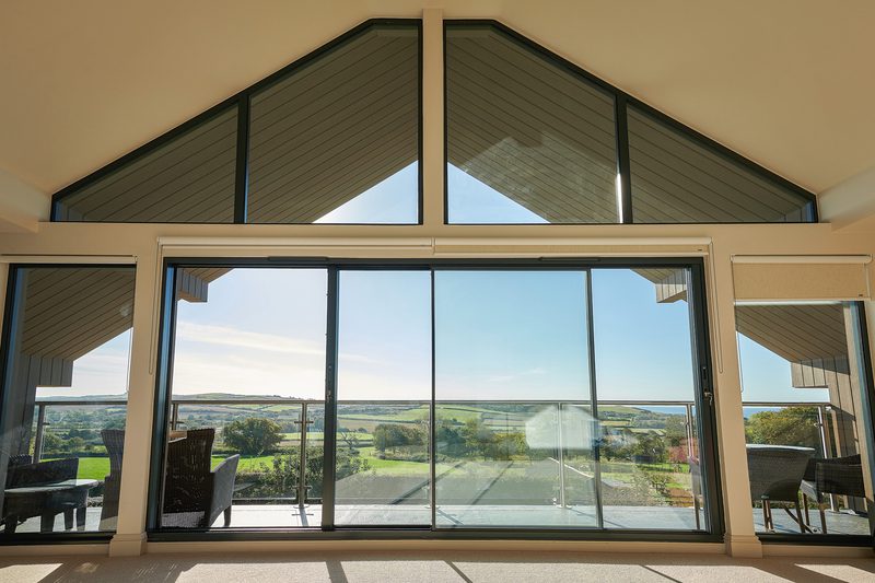 Bright living space with floor-to-ceiling glass doors opening to a balcony; triangular upper windows and a rural landscape of fields and trees beyond.