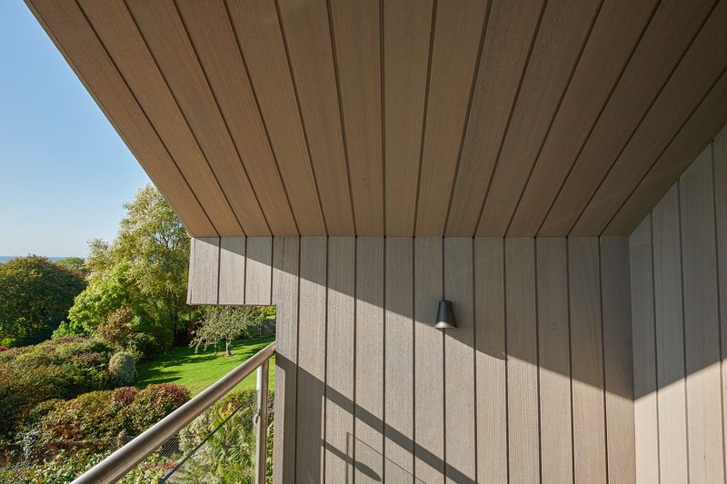 Balcony with beige wood paneling and slatted ceiling, glass railing, a wall light, and a sunlit garden with trees and a blue sky in the background.
