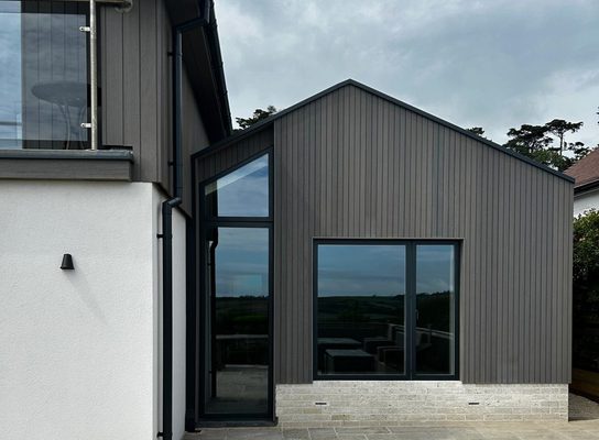 Modern house exterior with dark vertical siding, large glass doors and windows, a light brick base, white side wall, and a glass-railed balcony.
