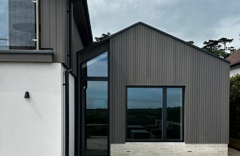Modern house exterior with dark vertical siding, large glass doors and windows, a light brick base, white side wall, and a glass-railed balcony.