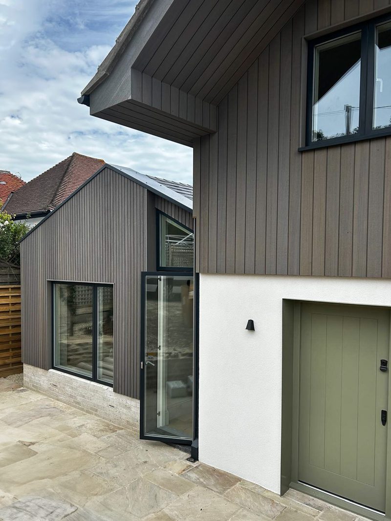 Modern house with dark vertical wood siding and white base, featuring a glass door and window, olive-green door, and a paved patio under a blue sky.