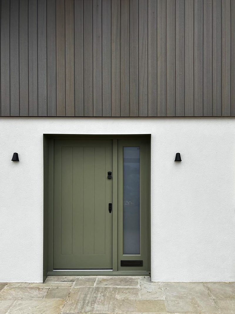 Olive green front door with a slim vertical glass panel on the right, flanked by white walls with two black exterior lights and a wood-clad facade.