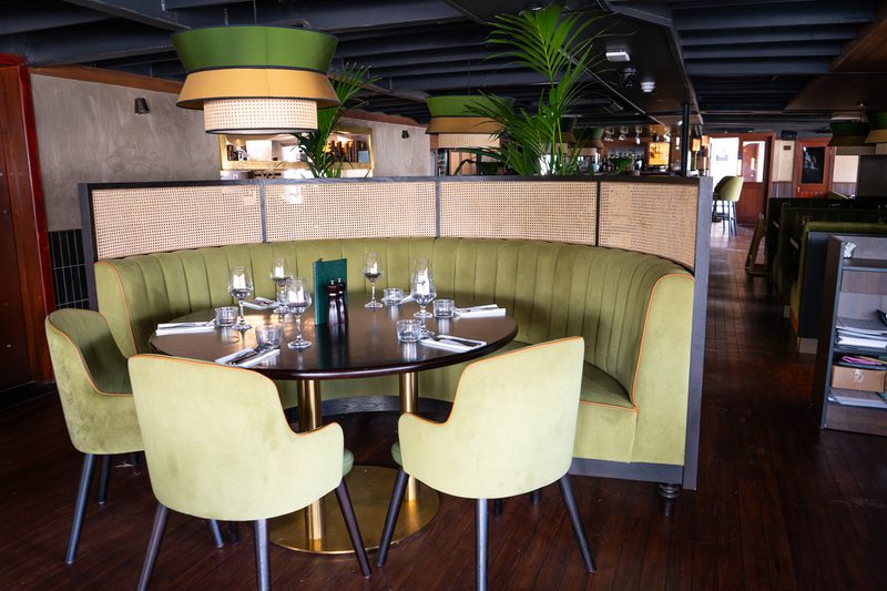 Round table surrounded by a curved olive-green booth and chairs in a stylish restaurant, set with glassware and cutlery, with pendant lights and plants.