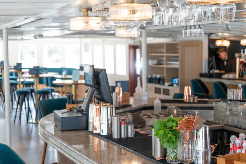 Bright, modern bar with a curved granite counter, copper barware, mint plant, and hanging glassware; blue-green stools and a bartender in back.
