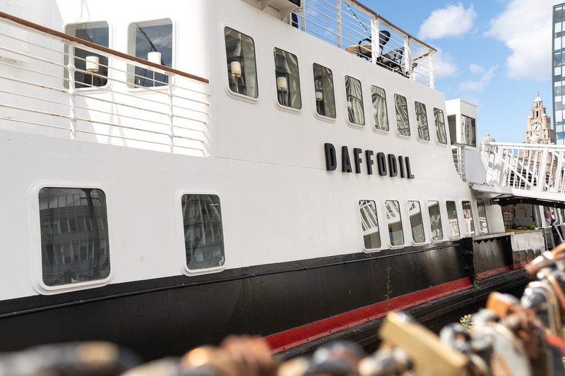 White passenger ferry named DAFFODIL with multiple windows, black hull with a red stripe, and upper-deck railings set against a blue sky with a distant cityscape.
