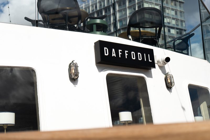 White boat exterior with a black rectangular sign reading DAFFODIL, round cabin windows, two vintage lanterns, and city buildings reflected in the glass.