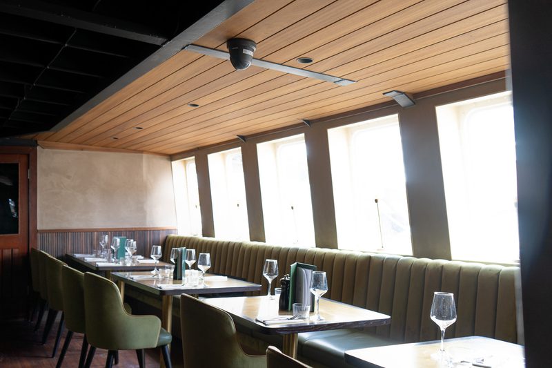 Restaurant dining area with a long olive-green banquette and chairs, tables set with wine glasses and menus, bathed in natural light from tall windows.